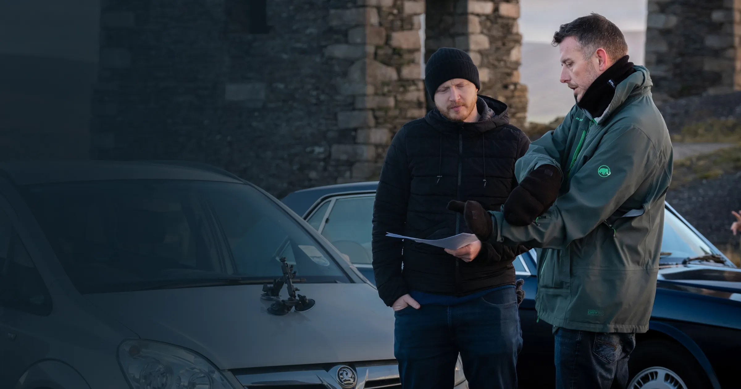 Two men stand by a silver car with a clipboard, wearing winter clothing. Stone ruins are in the background under a moody, cloudy sky.